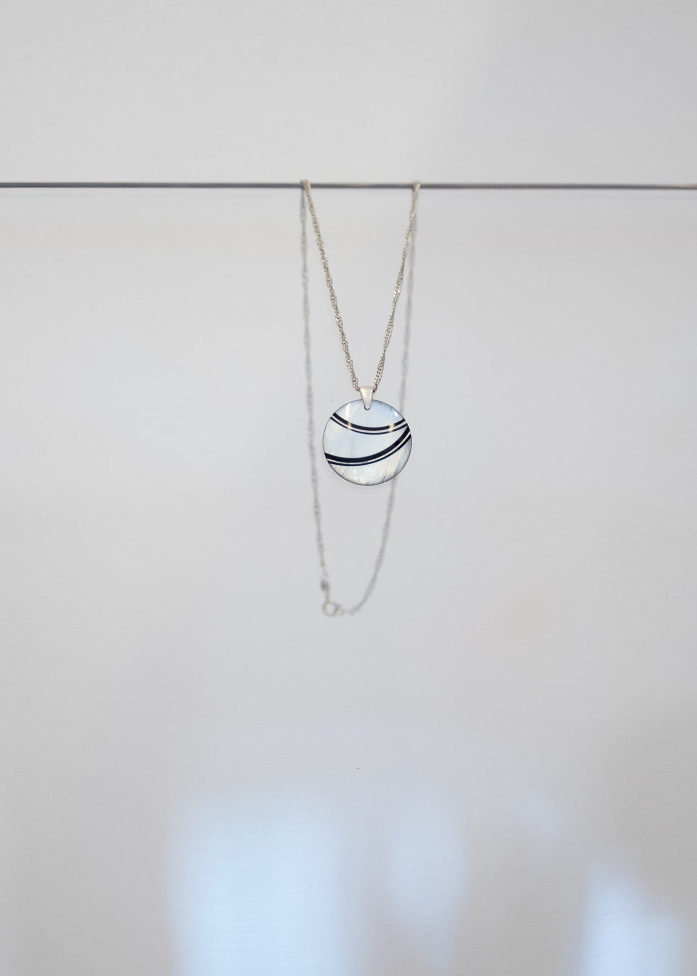 Collier « Onde » — pendentif rond en nacre blanche traversé de deux courbes noires, sur chaîne en argent torsadée. Joyeux Joyaux par Alexandra Lambert.